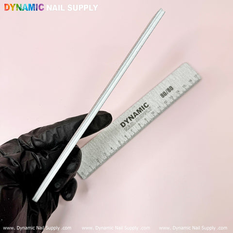 A gloved hand elegantly holds a square, white nail file vertically against a pink background, making it perfect for manicure enthusiasts. Next to it is a silver nail file labeled with the Dynamic Nail Supply brand, featuring the "Strong Emery Board - Professional Acrylic Nail Files (25 pcs)" in 100/180 grit—ideal for shaping acrylic nails. The image showcases the Dynamic Nail Supply logo prominently along with their website URL at both the top and bottom edges.