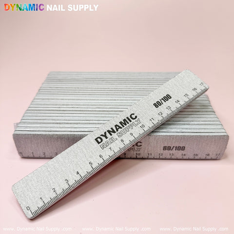 A stack of rectangular nail files is showcased on a light pink surface. A reusable nail file sits on top, featuring the Strong and Hard professional Jumbo nail files label by Dynamic Nail Supply with grit numbers 80/100 clearly visible. The brand name is printed in colorful letters at the top left corner on a pink background.
