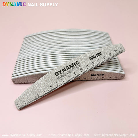 A stack of Professional Jumbo Half-moon Nail Files from Dynamic Nail Supply, ideal for acrylic nails, is displayed on a light pink background. One reusable nail file leans prominently at the front, showcasing the "DYNAMIC NAIL SUPPLY" brand and "100/180" grit specifications. A ruler edge can be seen, with the Dynamic Nail Supply logo elegantly positioned in the top left corner.