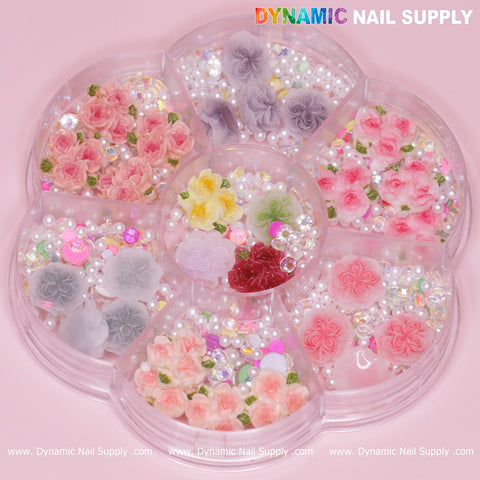 A container shaped like a flower holds a selection of colorful nail art decorations, including 40 pieces of white and pink 3D and 5D rose flower charms with pearl accessories. The lid features the Dynamic Nail Supply brand name. This set is perfect for creating Valentine nails art designs against a pink background.