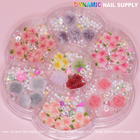 A flower-shaped container holds a stunning collection of vibrant 3D and 5D rose flower charms in pink, yellow, red, and gray, beautifully accented with pearls and rhinestones. The Dynamic Nail Supply brand name elegantly frames the top and bottom edges on a soft pink background, making it perfect for Valentine's nails art design.