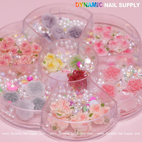 A round plastic storage container with various sections contains an assortment of nail art decorations, featuring white and pink 3D and 5D rose flower charms, pearls, and vibrant rhinestones—ideal for crafting Valentine nail art designs. The name Dynamic Nail Supply appears on the image.