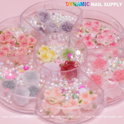 A circular container with multiple compartments showcases the 40 pcs White and Pink 3D 5D Rose Flower Charm with pearls, perfect for Valentine nails art designs. The sections contain pastel-colored roses, pearls, and sparkling gems. The brand name, Dynamic Nail Supply, is prominently displayed at the top. A pink background frames this elegant display, with the website URL repeated at the bottom.