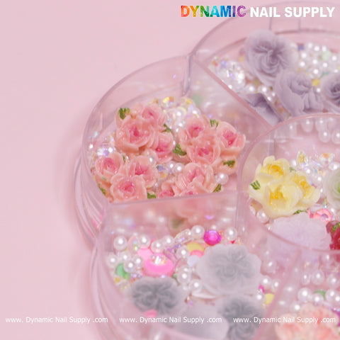 The Dynamic Nail Supply 40 pcs set of white and pink 3D and 5D Rose Flower Charms with pearls is displayed in a round nail art container. This container, featuring various compartments on a pink background, is perfect for Valentine Nails Art Design. Included are pink, yellow, and gray flower decorations alongside white pearls and multicolored gems. The DYNAMIC NAIL SUPPLY logo can be seen prominently in the corner.