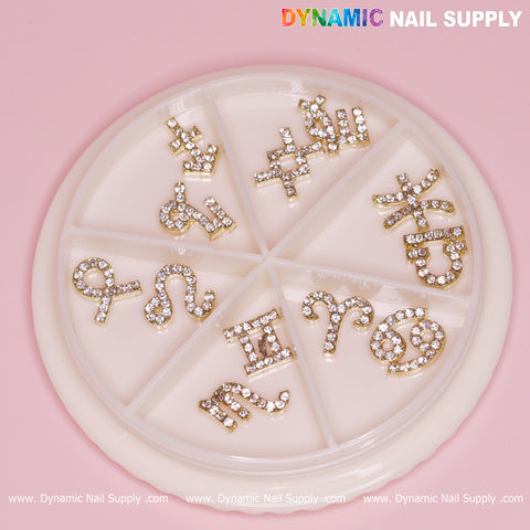 A circular nail art wheel with compartments displays a set of 12 Gold Zodiac Sign Nail Charms with rhinestone embellishments from Dynamic Nail Supply against a pink background. The colorful branding at the top reads "Dynamic Nail Supply," showcasing symbols for Aries, Taurus, Gemini, Cancer, Leo, Virgo, Libra, Scorpio, Sagittarius, Capricorn, Aquarius, and Pisces.
