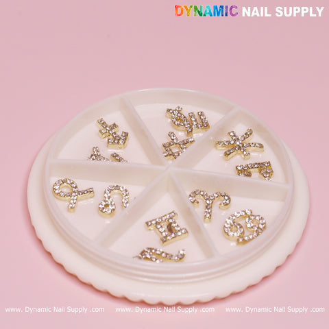 A round white container with eight compartments holds a Set of 12 pcs Gold Zodiac Sign Nails Charm with Rhinestone (Zodiac Symbol) from Dynamic Nail Supply. The background is a soft pink, and the brand name is printed at the top in colorful text.