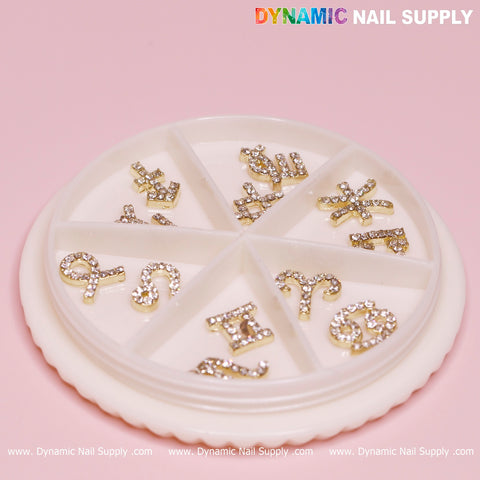 A round organizer featuring gold zodiac sign nail charms adorned with rhinestones, each in separate compartments. The background is light pink, showcasing the DYNAMIC NAIL SUPPLY logo in vibrant text at the top right corner. The organizer sits on a white surface, with the brand's website subtly repeated at the bottom.