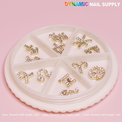 A round white container featuring seven compartments, each containing gold zodiac sign nail charms with rhinestones set against a pink background. The brand name Dynamic Nail Supply is showcased in colorful and white text at the top and repeated in white along the bottom.