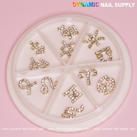 Round container featuring a set of 12 gold zodiac nail charms, each beautifully accented with rhinestones. The symbols—Scorpio, Sagittarius, Capricorn, Aquarius, Pisces, Aries, Taurus, and Gemini—glimmer against a soft pink backdrop. Brand name "Dynamic Nail Supply" prominently displayed at the top.