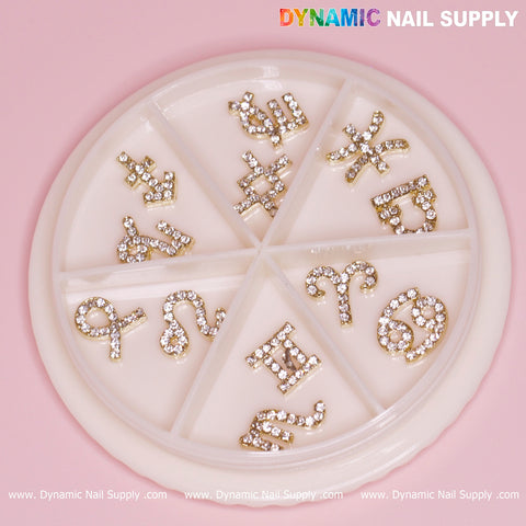 On a pink background, a circular container displays twelve compartments, each containing shimmering nail charms in the shape of gold zodiac signs adorned with rhinestones. At the top, "Dynamic Nail Supply" is printed in vibrant text.