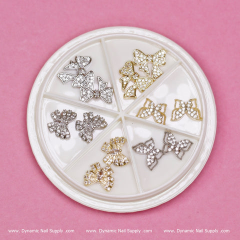 Set against a pink background, a circular white tray from Dynamic Nail Supply showcases six sets of metallic 3D butterfly-shaped nail charms. Each set glistens with tones of silver, gold, or rose gold and is adorned with small rhinestones—perfect for any Nail Art Designer in search of sophisticated embellishments.