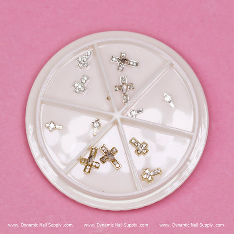 A round white container showcases segmented sections filled with eye-catching 3D Crosses Charms for Halloween nail art design (style #2) from Dynamic Nail Supply. The charms, available in both silver and gold, are embellished with tiny rhinestones set against a pink background. The text "Dynamic Nail Supply" is repeated at the bottom.