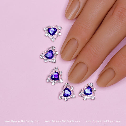 A hand with nude-colored nails rests on a pink surface. Alongside the nails, five heart-shaped charms featuring blue stones, part of Dynamic Nail Supply's "20 pcs Heart shape Charm for Nails Art Design," are arranged parallel to the fingers, highlighting an exquisite nail art design. The background includes a subtle website watermark.