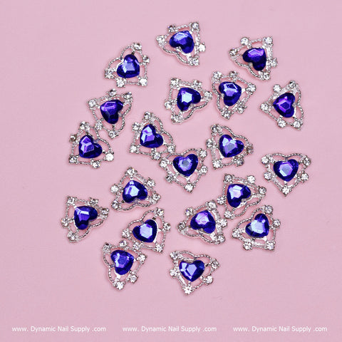 A display of nail art charms on a pink background features heart-shaped pieces embellished with blue gems, each surrounded by small, clear rhinestones arranged in a triangular pattern. The scattered design radiates elegance, while the Dynamic Nail Supply logo is prominently placed in the bottom corner.