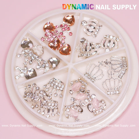 The Dynamic Nail Supply brand offers a round nail art wheel that includes 24 pieces of rhinestone-engraved Luxury 3D Crown Charms, ideal for Valentine's nail designs. This collection features pink hearts and ornate silver motifs, all beautifully displayed against a soft pink background.