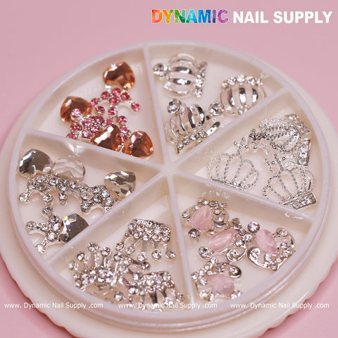 The 24-piece set of Luxury 3D Crown Charms from Dynamic Nail Supply showcases a romantic aesthetic, featuring rhinestone-engraved crowns, hearts, and clusters in elegant silver and pink shades. This collection is perfect for Valentines nail designs, packaged in a circular wheel with six compartments. The Dynamic Nail Supply logo is prominently displayed on the top against a pink background.