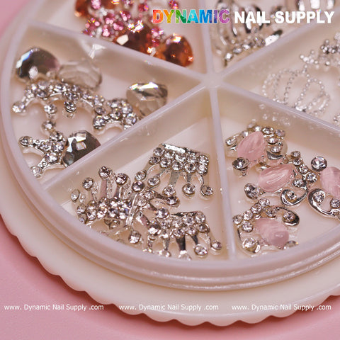 A circular organizer with six compartments contains a variety of dazzling nail art decorations, such as silver gemstones, pink stones, and intricate designs. The 24 pcs Luxury 3D Crown Charms (Rhinestone engraved) for Valentines Nail Designs add an elegant touch against the soft pink background. The brand name "Dynamic Nail Supply" is prominently displayed at the top and bottom.