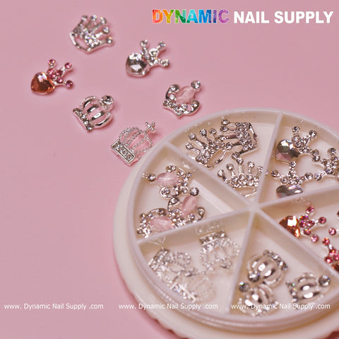 A pink background showcases assorted decorative nail gems, ideal for Valentine's Nail Designs, accompanied by a round plastic wheel container. The collection includes Luxury 3D Crown Charms and features heart and teardrop shapes in silver and pink hues. Text at the top reads Dynamic Nail Supply.