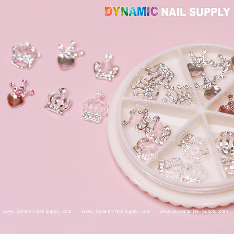 A nail decoration set featuring 24 pieces of Luxury 3D Crown Charms with rhinestone engravings, perfect for Valentine's nail designs. The set is neatly arranged in a round, sectioned container. Additional loose decorations in silver and pink colors are displayed on the pink surface, with the Dynamic Nail Supply branding clearly visible.