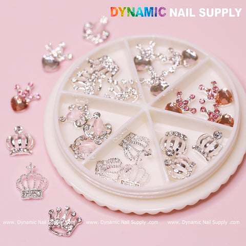 A round container with a pink background houses the 24 pcs Luxury 3D Crown Charms, specifically designed for Valentine's nail art, featuring intricate silver crowns and pink hearts adorned with shimmering rhinestones. Some decorations are artistically displayed outside the container, while the brand name "Dynamic Nail Supply" is prominently visible at the top.
