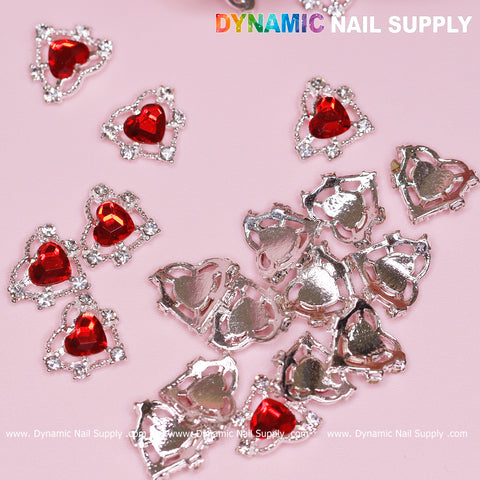 A close-up of the metallic heart-shaped nail charms from Dynamic Nail Supply displays their red gemstone centers scattered across a pink surface. Some charms are positioned upright, highlighting the vibrant red stones and silver edges, while others reveal their simple silver backs. These 20 pcs 3D Heart rhinestone Charms (Red stones + Silver claws base) are perfect for your next nail art design project.