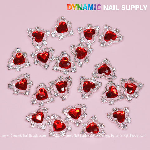 A set of 20 pieces of 3D heart rhinestone charms for nail art design, featuring red stones with a silver claw base, is elegantly displayed on a pink surface. The top is adorned with the vibrant logo of Dynamic Nail Supply. These scattered nail charms highlight their dazzling design and distinct nail art appeal.