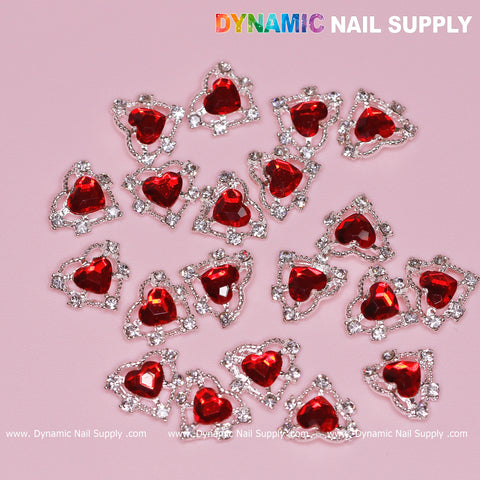 Red heart-shaped rhinestones encased in silver claws are set on a pink background. The casually arranged stones from the "20 pcs 3D Heart Rhinestone Charm for Nails Art Design (Red stones + Silver claws base)" create an elegant nail art design with "Dynamic Nail Supply" prominently displayed in white at the top, and the website URL forming a decorative pattern at the bottom.