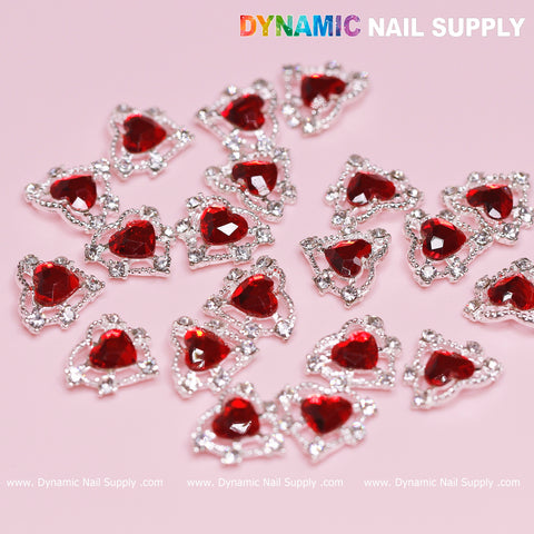 A set of 3D heart-shaped rhinestones, featuring red stones with a silver claw base, is displayed on a light pink surface. The top right corner showcases the text Dynamic Nail Supply. These dazzling and reflective charms are ideal for elevating any nail art design or decoration.