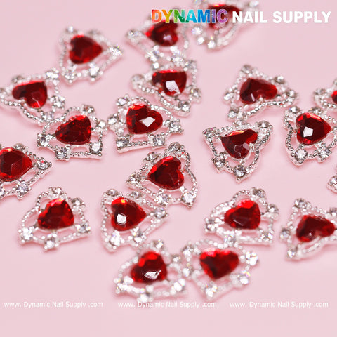 A set of 20 pieces of 3D heart-shaped rhinestone charms for nail art design, featuring vibrant red stones with a silver claw base, are artfully arranged on a light pink background. The intricate silver detailing gives each stone a jewelry-like appearance. At the top, in bold letters, is the name "Dynamic Nail Supply.