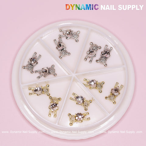 Displayed against a light pink background, the Dynamic Nail Supply product features a round white container containing six compartments. It includes 12 pieces of metallic dog-shaped and teddy bear charms with engraved rhinestones. Half of the sections contain gold heart-shaped rhinestones, while the other half hold similarly designed silver ones. The brand name, Dynamic Nail Supply, is prominently placed in the upper right corner.