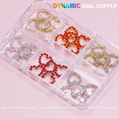 A clear plastic container with six compartments contains decorative nail embellishments in the shape of open hearts. These embellishments feature gold, red, and silver hues embellished with sparkling rhinestones, making them perfect for elevating any nail art design. The brand "Dynamic Nail Supply" is displayed in the top right corner against a pink background.