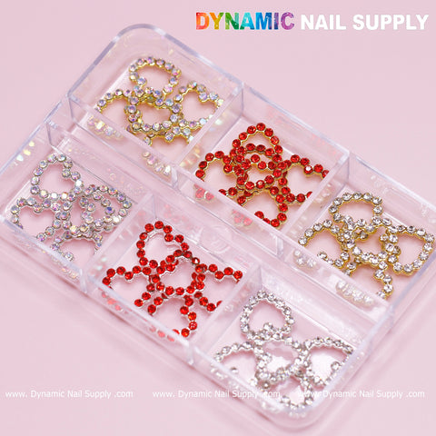 A clear plastic organizer features six compartments filled with heart-shaped nail decorations, ideal for any nail art design. These compartments contain rhinestones in red, silver, and gold colors. The brand name "Dynamic Nail Supply" is printed at the top in colorful letters against a soft pink background.