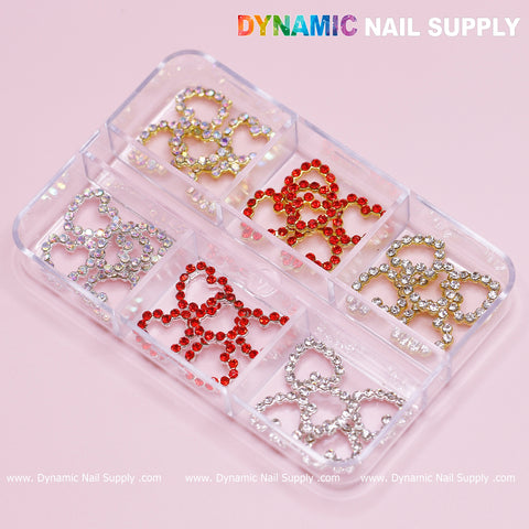 A clear plastic container featuring six compartments contains an assortment of decorative nail art pieces, including metallic bow-shaped embellishments in silver, red, and gold. Ideal for any nail art design, the container is set against a pink background with "Dynamic Nail Supply" artistically printed above it.