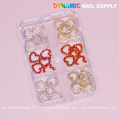 A clear plastic case holds sparkling heart-shaped rhinestone charms bordered with rhinestones in three colors: silver, gold, and red. Each compartment contains identical open heart charms neatly arranged, ideal for any nail art design. The pink background showcases the brand name Dynamic Nail Supply in colorful lettering at the top.