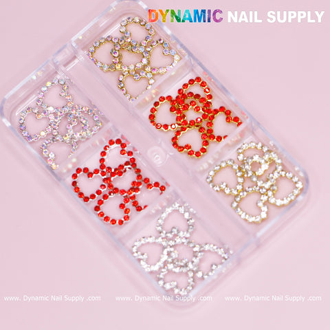 A transparent plastic container features six compartments, each filled with decorative heart-shaped pieces perfect for nail art. These 30 pcs Rhinestones Heart Charm for Nails Art Design boast open heart shapes outlined in silver, gold, and red borders. The brand name Dynamic Nail Supply is prominently displayed at the top right corner in vibrant text against a pink background, making it ideal for your next nail design endeavor.