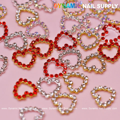 Scattered across a light pink surface, the 30 pcs Rhinestones Heart Charm for Nails Art Design in red, gold, and silver create an exquisite display. With delicate patterns crafted from rhinestones with a border edge and open heart design, Dynamic Nail Supply is prominently featured in this colorful and decorative composition.
