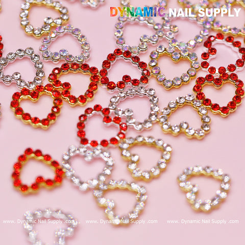 A pink background is adorned with a dazzling collection of 30 pcs heart-shaped rhinestones, ideal for nail art design. These charms, available in red, silver, and gold with a rhinestone border edge and open heart design, sparkle brilliantly. "Dynamic Nail Supply" is prominently displayed at the top in vibrant letters, while the website URL is elegantly repeated at the image's bottom.