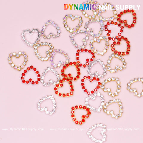 A scattering of 30 pieces of Rhinestones Heart Charm for Nails Art Design featuring red, gold, and silver charms on a pink background creates a dazzling effect. The sparkling appearance captivates with its Rhinestones Border Edge Open Heart design. Dynamic Nail Supply, renowned for its innovative nail art designs, is prominently displayed at the top in colorful letters.