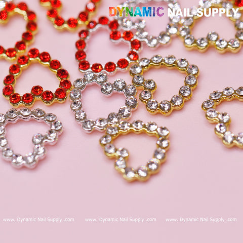 A pink surface is adorned with 30 chic heart-shaped rhinestone charms in red, silver, and gold, ideal for enhancing your Nails Art Design. Casually arranged for ultimate appeal, these stylish charms exemplify the allure of Dynamic Nail Supply. The top features the text "DYNAMIC NAIL SUPPLY," accompanied by a repeating website URL below.