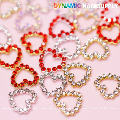A delightful assortment of 30 Rhinestones Heart Charms from Dynamic Nail Supply, showcasing red, silver, and gold rhinestones with a border edge and open heart design, is elegantly arranged on a pink background. The hollow centers of the hearts enhance this nail art design. "Dynamic Nail Supply" is displayed in multicolored letters at the top left, while "www.DynamicNailSupply.com" graces the bottom edge.
