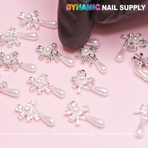 A gloved hand arranges the 20 pcs Silver (Ribbon) Bow shape Dangle Charms with pearl for nails on a pink surface. The charms, featuring pearl-like beads and rhinestones in elegant silver bow shapes adorned with teardrops, shine brilliantly. "Dynamic Nail Supply" is prominently displayed at the top, subtly echoed at the bottom.