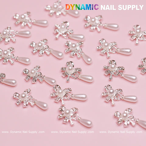 Close-up of decorative nail charms arranged on a pink surface, showcasing the 20 pcs Silver (Ribbon) Bow shape Dangle Charms with pearls for nails. The top of the image displays the text Dynamic Nail Supply in colorful letters. Perfect for any nail designer seeking unique flair.