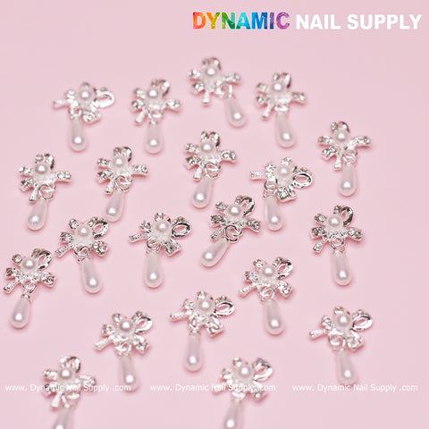 A collection of 20 silver bow-shaped dangle charms, each featuring a central pearl, is elegantly arranged on a pink background. These charms, crafted by Dynamic Nail Supply, embody the refined touch of an expert nail designer.