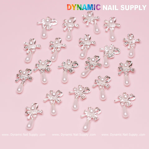 The image features a variety of silver bow-shaped nail art dangle charms, each with a sophisticated design containing a pearly centerpiece and a dangling teardrop-shaped pearl. These elegant nail accessories are arranged on a pink background, with "Dynamic Nail Supply" written in white at both the top and bottom.