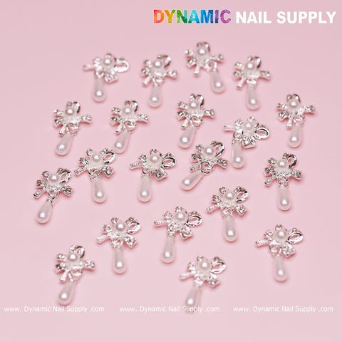 A set of 20 silver ribbon bow-shaped dangle charms is elegantly displayed against a pink background. Each charm features a small pearl and floral rhinestone design, complemented by a teardrop-shaped pearl hanging below. The top right corner displays the "Dynamic Nail Supply" brand in vibrant, colorful letters.