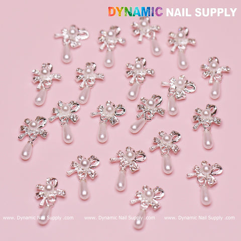 Various silver bow-shaped dangle charms with pearls from the "20 pcs Silver (Ribbon) Bow shape Dangle Charms with pearl for Nails Designer" collection by Dynamic Nail Supply are scattered across a light pink surface. Each charm includes small pearls, rhinestones, and a dangling pearl-like bead. The brand name Dynamic Nail Supply is prominently displayed at the top, and the website is repeated at the bottom.