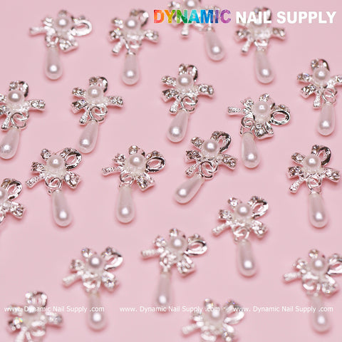 The 20-piece set of silver bow-shaped dangle charms with pearl accents for nails is displayed against a pink background. The top right corner features the text Dynamic Nail Supply. These designer charms showcase shiny ribbon designs and dangling pearl-like teardrops, giving them an elegant appearance.
