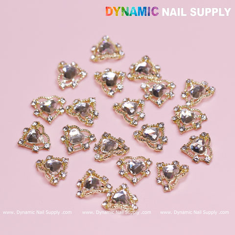 A set of 20 luxurious heart charms featuring rhinestone border edges with gold trim, beautifully arranged on a pink background. These sparkling stones are intricately designed with small surrounding gems, making them perfect for Valentine nail designs. The brand "Dynamic Nail Supply" is displayed in multicolored and white text at the top right and bottom center.