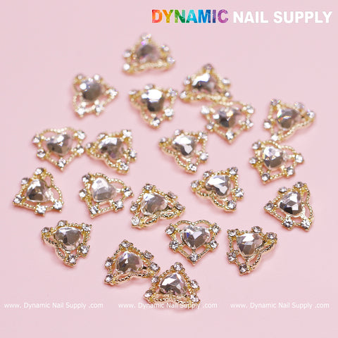The 20 pcs Luxury Heart Charm for Valentine Nails Design from Dynamic Nail Supply features decorative heart and triangle charms adorned with a clear central gem surrounded by small rhinestones set in gold. These charms, perfect for Valentine's nail designs, are displayed on a light pink background with the Dynamic Nail Supply logo in the top right corner.