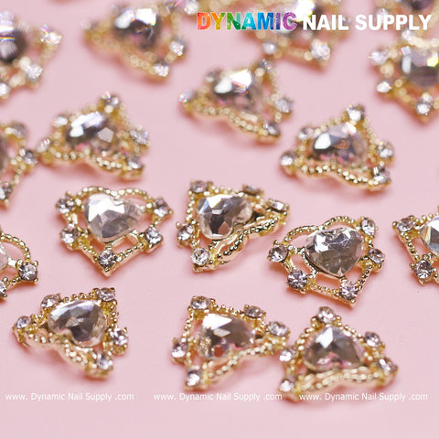 A close-up shot features luxury heart-shaped and triangular gold nail charms with clear gemstones, artfully arranged on a pink background. The rhinestones border edge enhances their elegance. At the top, the image displays the text Dynamic Nail Supply, with their website URL at the bottom. This set of 20 pcs Luxury Heart Charm by Dynamic Nail Supply is ideal for your Valentine nails design.